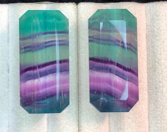 May include: Two rectangular fluorite gemstones with emerald green, purple, and clear bands. The gemstones have a faceted cut and are displayed on a white surface. The stones are polished and reflect light.
