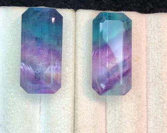 May include: Two rectangular, faceted gemstones with a mix of blue, teal, and purple hues. The stones are displayed on a white surface against a dark background. The gems have a polished, translucent appearance.