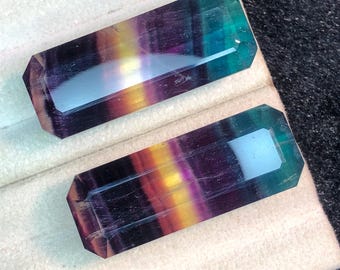 Könnte beinhalten: Zwei rechteckige Fluorite-Edelsteine. Die Steine zeigen einen Farbverlauf aus Lila, Gelb und Grün. Die Edelsteine sind facettiert mit einer polierten Oberfläche und werden auf einer weißen Oberfläche präsentiert.