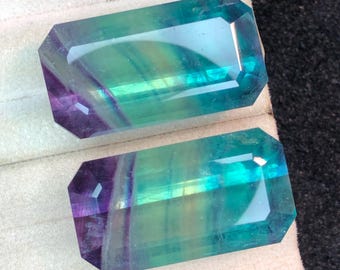 May include: Two rectangular fluorite gemstones. The gemstones have a mix of colours, including purple, green, and blue. The gemstones are faceted with a rectangular shape and bevelled edges. The gemstones are displayed on a white surface.