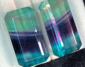 May include: Two rectangular, faceted gemstones with a vibrant color gradient. The stones transition from teal to purple and green, with a polished, reflective surface. The gems are displayed on a soft, white surface.