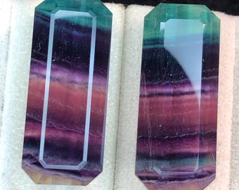 May include: Two rectangular fluorite crystals with a mix of purple, green, and yellow hues. The crystals have a faceted, geometric design and are displayed on a white surface. The gemstones have a polished, translucent appearance.