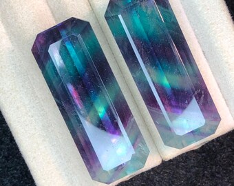 May include: Two rectangular fluorite crystals with a mix of purple, teal, and blue hues. The crystals are faceted, showcasing their internal colors and clarity. They are displayed on a white and gray surface.