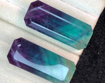 May include: Two rectangular fluorite crystals with a polished, faceted design. The crystals display a gradient of colors, including purple, teal, and green. The crystals are displayed on a white, textured surface.