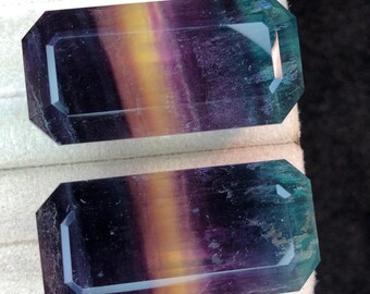 May include: Two rectangular gemstones with a multi-coloured pattern of purple, yellow, and teal. The gemstones have a faceted edge and are displayed on a white surface.