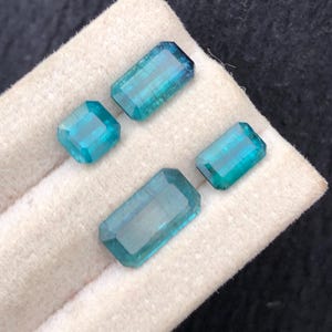 Puede incluir: Cuatro piedras preciosas de color turquesa, talladas en forma rectangular y cuadrada, se presentan sobre una tela crema. Las gemas son transparentes, con facetas que reflejan la luz. La disposición resalta los diferentes tamaños y formas, para joyería. Medidas en cm.