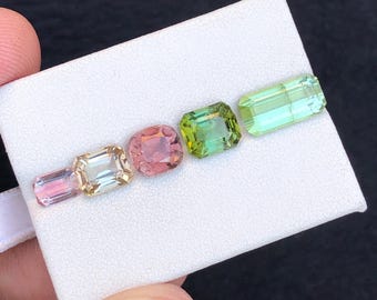 May include: A collection of six colorful gemstones in various shapes and hues, including pink, yellow, green, and blue-green. The gems are arranged on a white surface, showcasing their facets and clarity. The gemstones are likely for jewelry making.