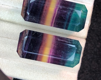 May include: Two rectangular fluorite gemstones in shades of purple, yellow, and green. The gemstones have faceted edges and are presented on a white surface.