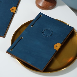 Può includere: Una copertina di menu in pelle blu con un logo in rilievo, fissata con un cordino nero e una linguetta angolare in pelle marrone chiaro. Il menu poggia su un vassoio color oro. Un portacandele in legno con una candela accesa è sullo sfondo.