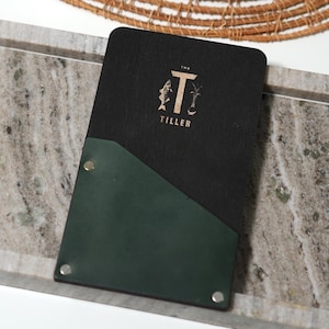 Puede incluir: Una cubierta de menú de madera negra con un bolsillo de cuero verde. La cubierta tiene un logotipo dorado que dice "The Tiller" con un pez estilizado y una palmera.
