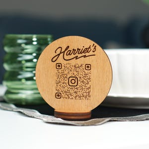Puede incluir: Un letrero redondo de madera con el nombre "Harriet's" en escritura cursiva y un código QR. El letrero está sobre una pequeña base de madera. Un vaso verde y un cuenco blanco están en el fondo.