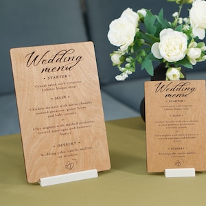 May include: Two wooden wedding menu signs on white stands. The menus are light brown with black text, including the words "Wedding menu" at the top. A bouquet of white flowers sits behind the signs. The menus list starter, main, and dessert options.