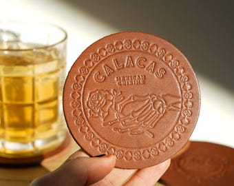 May include: A brown leather coaster with the word "CALACAS" embossed on it, along with the words "MEXICAN CUISINE" and a skeleton hand holding a rose. The coaster is round and has a decorative border.