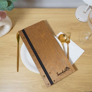 Può includere: Un menu in legno con la scritta "Sandals" è esposto su un tavolo, accanto a un piatto bianco, posate dorate e un tovagliolo bianco. Sono visibili anche un bicchiere da vino trasparente e un vaso decorativo con fiori rosa.
