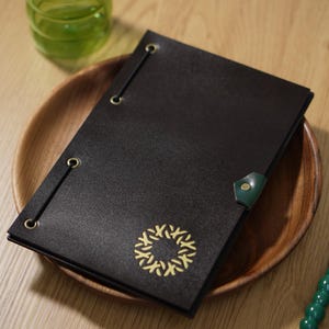 Puede incluir: Una cubierta de menú de cuero negro con un emblema dorado, asegurada con elásticos y ojales de latón. La cubierta está sobre un plato de madera, junto a un tenedor y un cuchillo con mangos verdes. Un vaso de líquido verde está en el fondo.