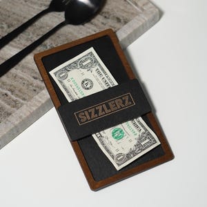 May include: A brown wooden check presenter with a black leather insert. The insert has the word "SIZZLERZ" printed in gold lettering. Two US dollar bills are placed inside the presenter.