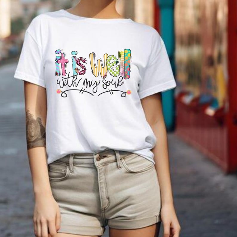 May include: White t-shirt with the phrase "it is well with my soul" in colorful, patterned letters. The design includes decorative swirls and dots. The shirt is short-sleeved.