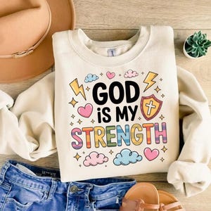 May include: Cream-colored sweatshirt with the text "GOD is MY STRENGTH" in colorful letters. The design includes hearts, clouds, stars, and lightning bolts. The sweatshirt is paired with blue jeans, a hat, and sandals.