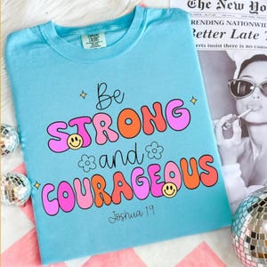May include: A light blue t-shirt with the words "Be STRONG and COURAGEOUS" in a retro font. The text is in pink and orange with smiley face and flower accents. The scripture reference "Joshua 19" is at the bottom.