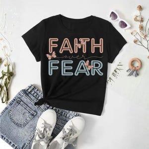 May include: Black t-shirt with the words "FAITH over FEAR" in a two-tone design, with butterfly accents. The shirt is paired with denim shorts and white sneakers, creating a casual, everyday look.