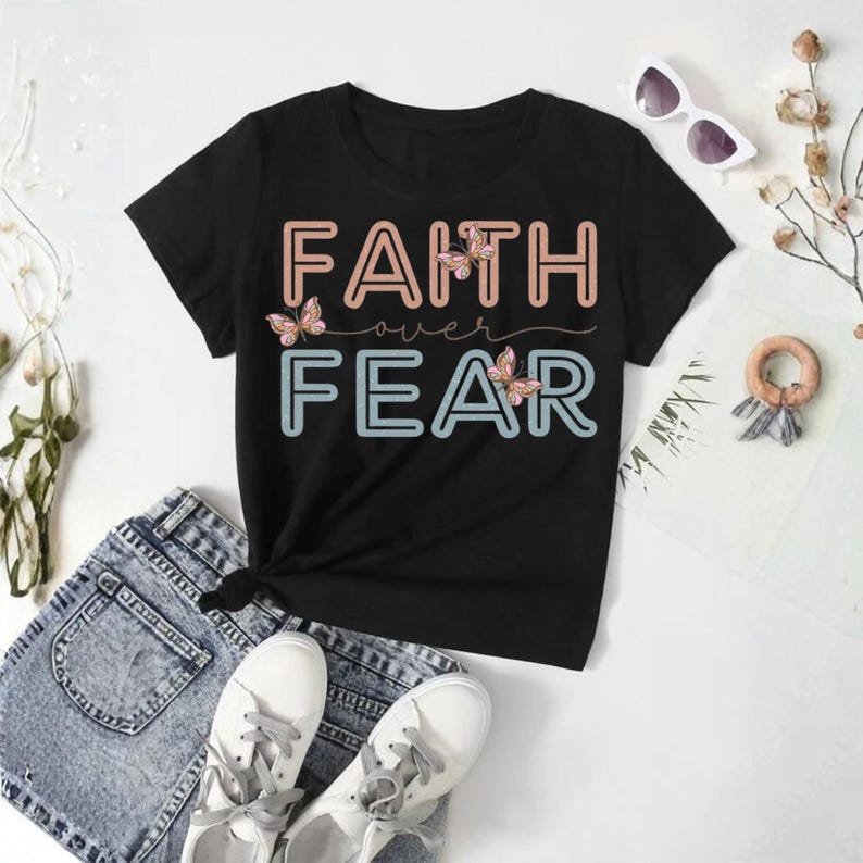 May include: Black t-shirt with the words "FAITH over FEAR" in a two-tone design, with butterfly accents. The shirt is paired with denim shorts and white sneakers, creating a casual, everyday look.