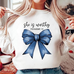 May include: White sweatshirt with a large blue bow graphic and the text "she is worthy PROVERBS 31" in black script. The bow is detailed with shading, giving it a 3D appearance. The sweatshirt is a casual, everyday garment.