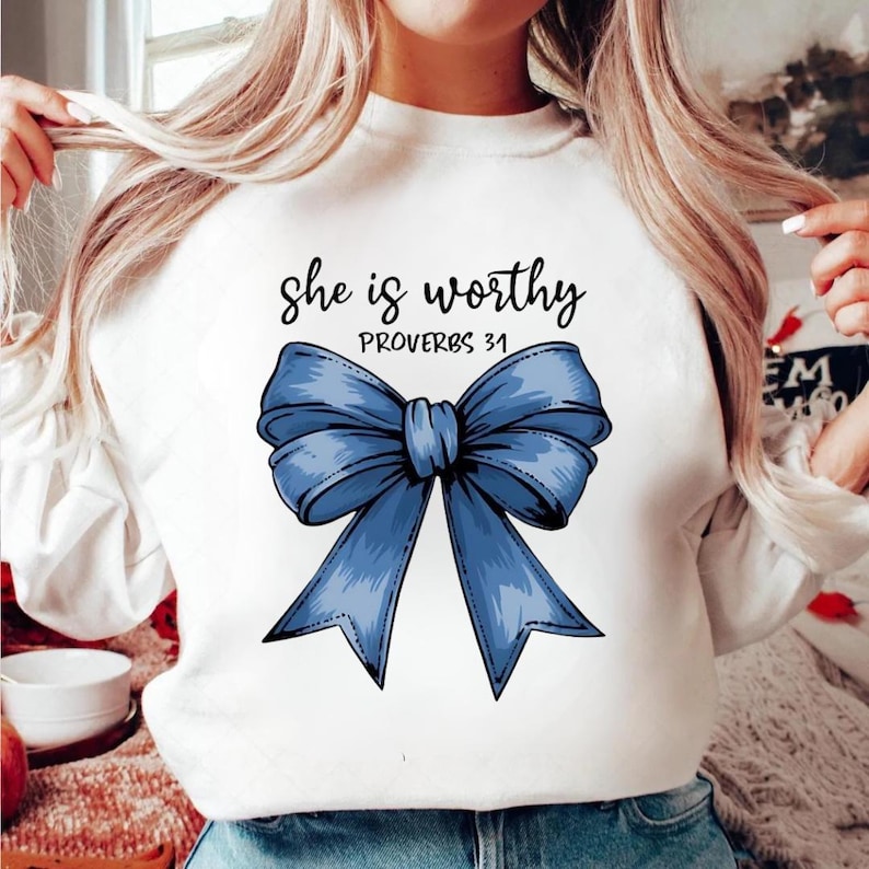 May include: White sweatshirt with a large blue bow graphic and the text "she is worthy PROVERBS 31" in black script. The bow is detailed with shading, giving it a 3D appearance. The sweatshirt is a casual, everyday garment.