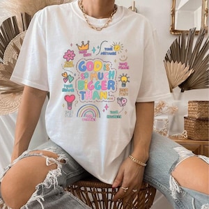 May include: White t-shirt with a colorful graphic that reads "GOD IS SO MUCH BIGGER THAN..." surrounded by various illustrations. The design includes a crown, sun, flower, heart, rainbow, and text like "Your Hurt" and "Your Fears".