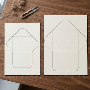 May include: Two off-white rectangular sheets of paper, each featuring a black printed outline of an envelope template with dashed fold lines. A pair of gold-toned metal scissors and a brown pen are positioned near the paper sheets.