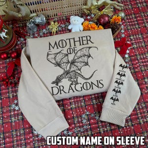 May include: Tan sweatshirt with the words "MOTHER OF DRAGONS" and a dragon graphic. The sleeve has custom names printed on it. The text "CUSTOM NAME ON SLEEVE" is at the bottom of the image.