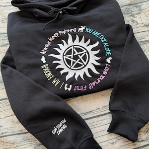 Może przedstawiać: Czarna bluza z kapturem z białym wzorem promieni słonecznych i pentagramem pośrodku. Frazy "Always Keep Fighting", "You Are Not Alone", "Love Yourself First" i "I Am Enough" otaczają wzór. Na rękawie widnieje napis "SW-DW-JW CASTIEL".