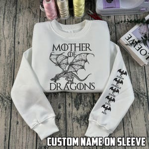 May include: White sweatshirt with the text "MOTHER OF DRAGONS" and a dragon graphic. The sleeve features custom names and dragon silhouettes. The phrase "CUSTOM NAME ON SLEEVE" is printed at the bottom.