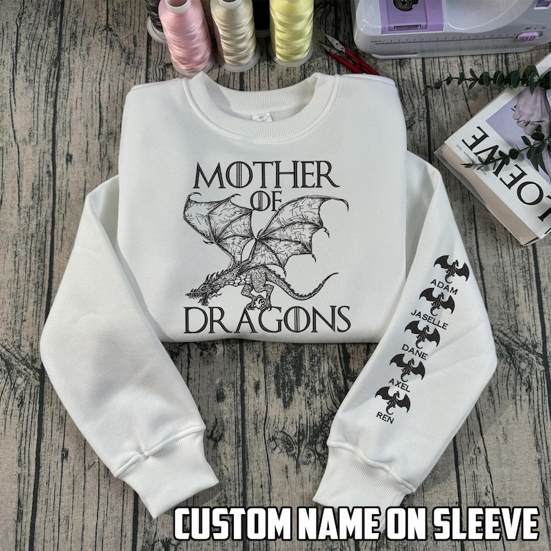 May include: White sweatshirt with the text "MOTHER OF DRAGONS" and a dragon graphic. The sleeve features custom names and dragon silhouettes. The phrase "CUSTOM NAME ON SLEEVE" is printed at the bottom.