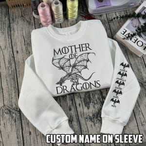 May include: White sweatshirt with the text "MOTHER OF DRAGONS" and a dragon graphic. The sleeve features custom names. The sweatshirt is made of a soft material and has a crew neck.