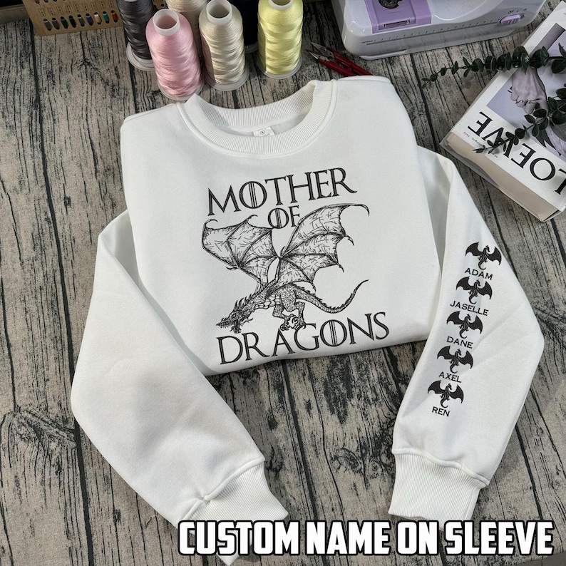 May include: White sweatshirt with the text "MOTHER OF DRAGONS" and a dragon graphic. The sleeve features custom names. The sweatshirt is made of a soft material and has a crew neck.