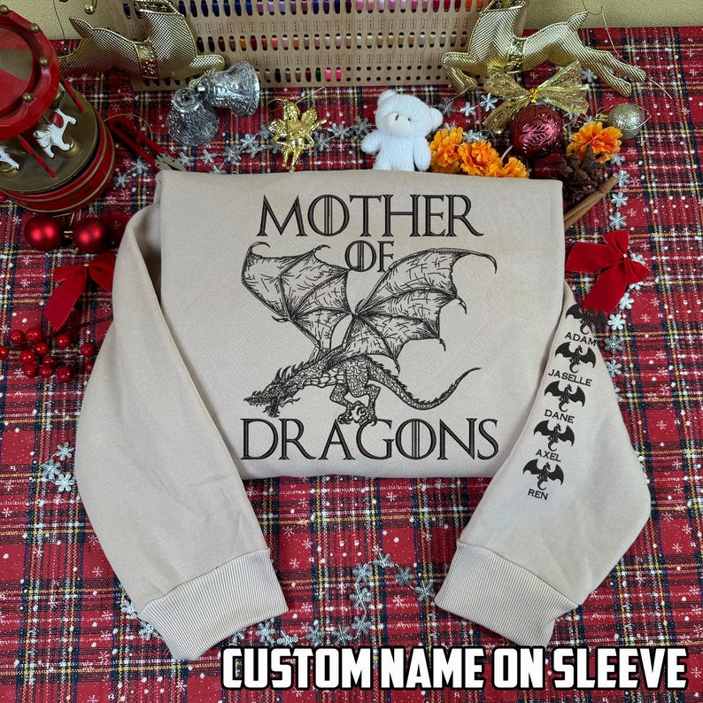 May include: A beige sweatshirt featuring a black dragon graphic with the words "MOTHER OF DRAGONS." The sleeve displays custom names. The text "CUSTOM NAME ON SLEEVE" is at the bottom. The sweatshirt is displayed on a red and green plaid surface.