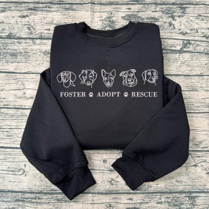 Peut inclure: Sweat-shirt noir avec un contour blanc de cinq têtes de chien et le texte "FOSTER ADOPT RESCUE".