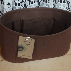 May include: A brown felt handbag organizer with multiple interior pockets. The organizer has an oval shape and a tag that reads "DIVIDE AND CONQUER". The product is designed to help organize items inside a handbag.