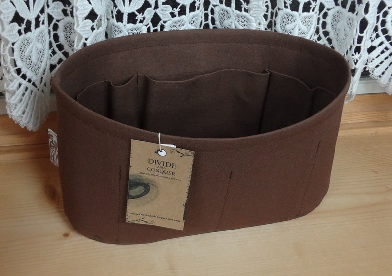 May include: A brown felt handbag organizer with multiple interior pockets. The organizer has an oval shape and a tag that reads "DIVIDE AND CONQUER". The product is designed to help organize items inside a handbag.