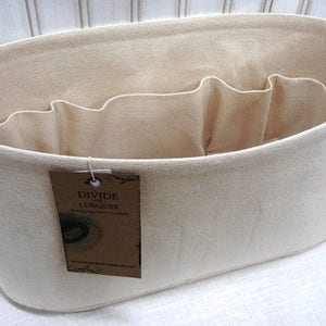 May include: A beige canvas tote bag with multiple interior compartments for organization. The bag has an oval shape and a tag that reads "DIVIDE and CONQUER". The bag is designed to help organize items.