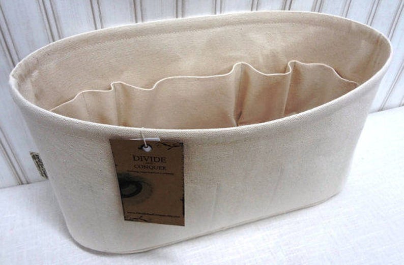 May include: A beige canvas tote bag with multiple interior compartments for organization. The bag has an oval shape and a tag that reads "DIVIDE and CONQUER". The bag is designed to help organize items.
