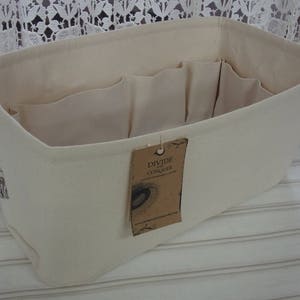 May include: Cream-colored rectangular organizer with multiple interior pockets. A brown tag with the words "DIVIDE CONQUER" hangs from the side. The organizer is made of a soft, neutral-toned fabric, ideal for storing items.