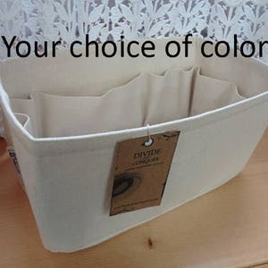May include: Off-white canvas organizer with multiple interior compartments. The text "Your choice of color" is displayed above the organizer. A brown tag with the text "DIVIDE CONQUER" hangs from the side.
