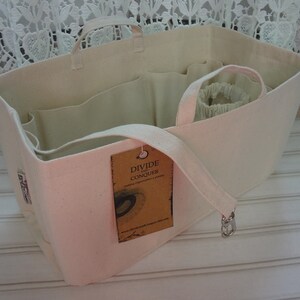 May include: A white canvas tote bag with multiple compartments and a zipper closure. The bag has a brown tag that reads "Divide and Conquer" and a white strap.