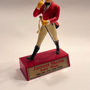 Könnte beinhalten: Figur des Johnnie Walker Scotch Whisky Striding Man. Die Figur trägt einen roten Mantel, einen gelben Zylinder und schwarze Stiefel und steht auf einem roten Sockel mit dem Markennamen und dem Text "Born 1820 - Still Going Strong".