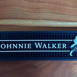 Może przedstawiać: Prostokątna czarna gumowa mata barowa o teksturowanej powierzchni z wypustkami. Mata zawiera biały napis "JOHNNIE WALKER" oraz białą grafikę przedstawiającą idącą postać w cylindrze, otoczoną dwiema poziomymi białymi liniami.