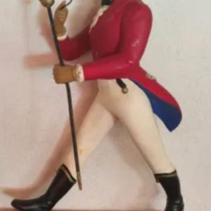 Op de afbeelding: Figuur van een man in een rode jas, beige hoge hoed en zwarte laarzen, met een wandelstok. De figuur staat op een rode basis met de gouden tekst "JOHNNIE WALKER".