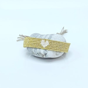 Peut inclure: Un bracelet en macramé doré avec un motif de cœur blanc. Le bracelet est enroulé autour d'une pierre grise. Les extrémités du bracelet ont des fils beiges.
