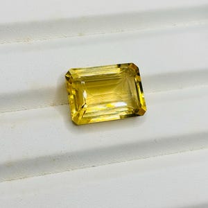 May include: A rectangular, faceted gemstone with a vibrant yellow hue. The gem has a clean, transparent appearance and is set against a white background. The cut of the stone allows light to reflect, creating a brilliant shine.