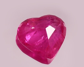 Puede incluir: Una gema de rubí facetada en forma de corazón. El rubí es de un color rosa intenso y vibrante con múltiples facetas que reflejan la luz. La gema está sobre un fondo rosa claro y suave.